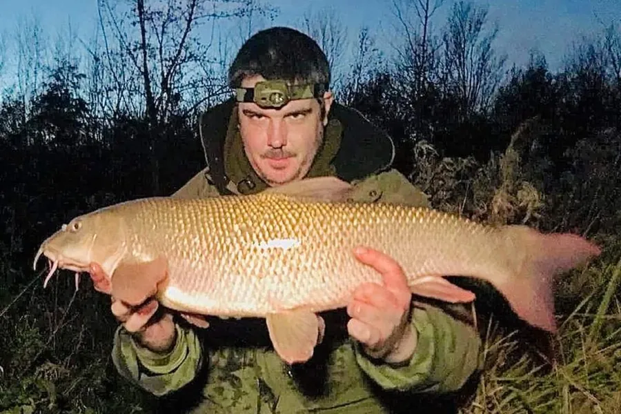 An angler at dusk holding a large barbel caught on a Nene backwater
